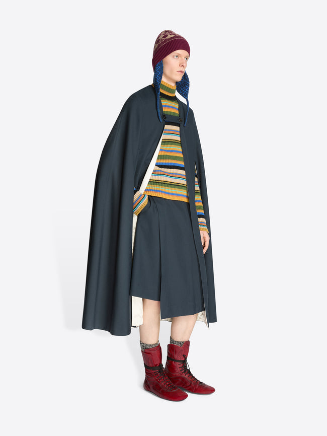 Person wearing a dark cape, striped sweater, skirt, hat and red boots on a white background