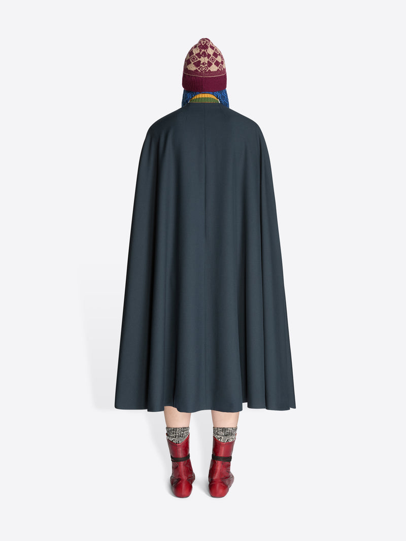 Back view of a person wearing a dark cape, striped sweater, skirt, hat and red boots on a white background
