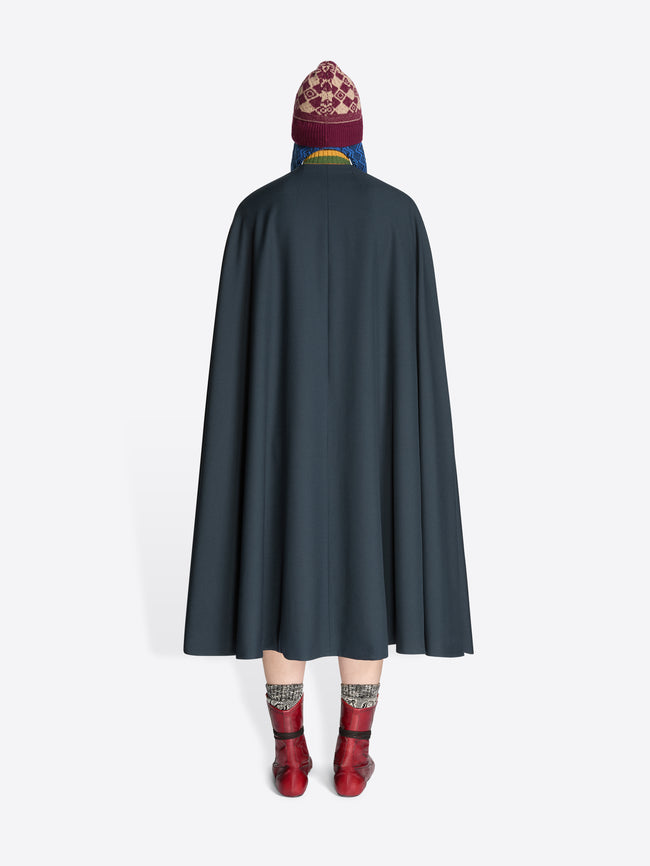 Back view of a person wearing a dark cape, striped sweater, skirt, hat and red boots on a white background