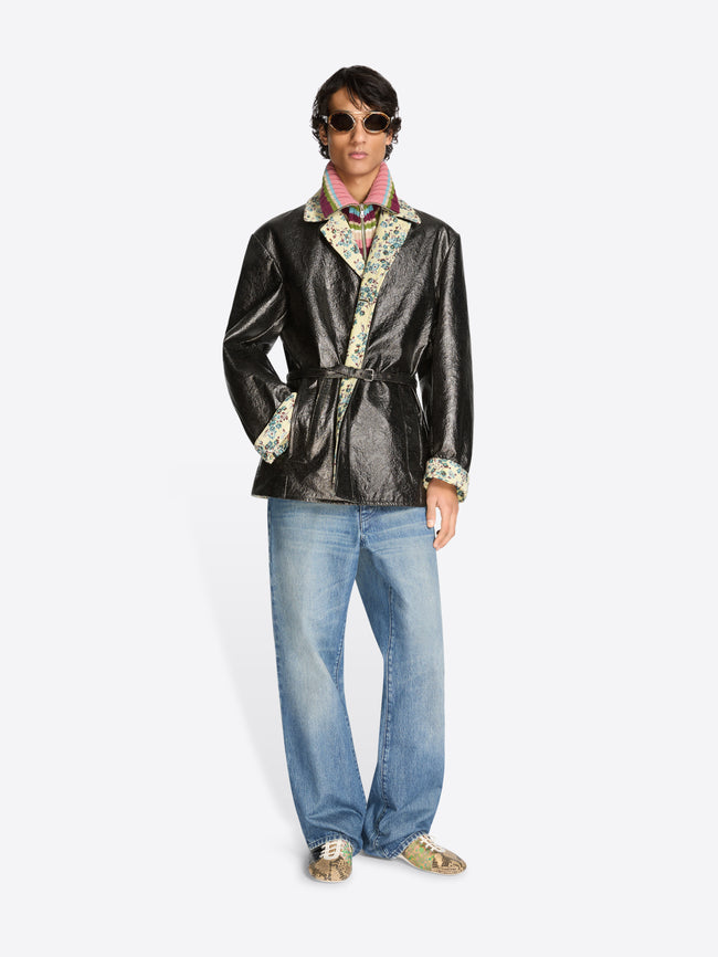 Person wearing a shiny black coat with floral patterns, blue jeans, and shoes on a white background