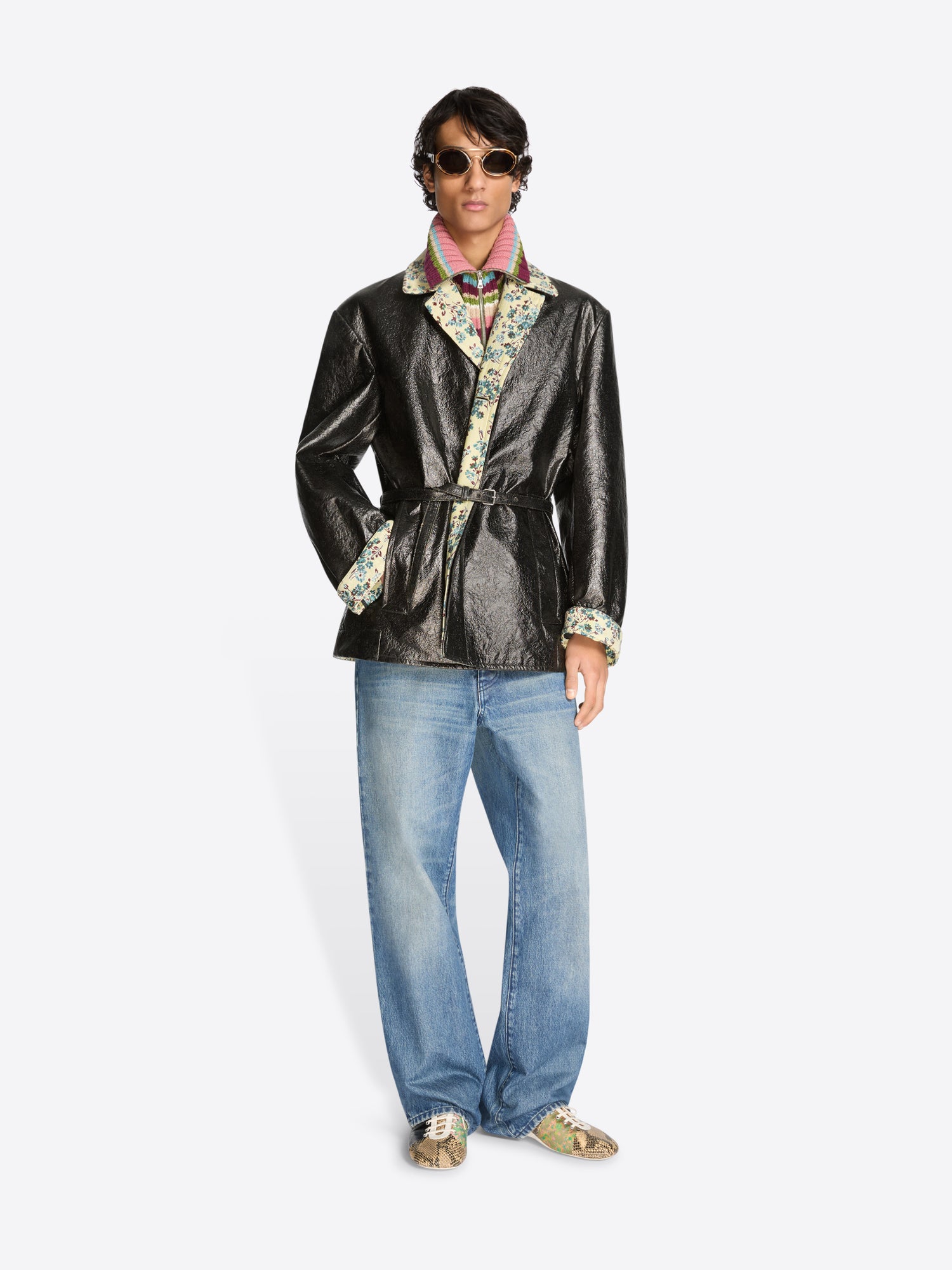 Person wearing a shiny black coat with floral patterns, blue jeans, and shoes on a white background