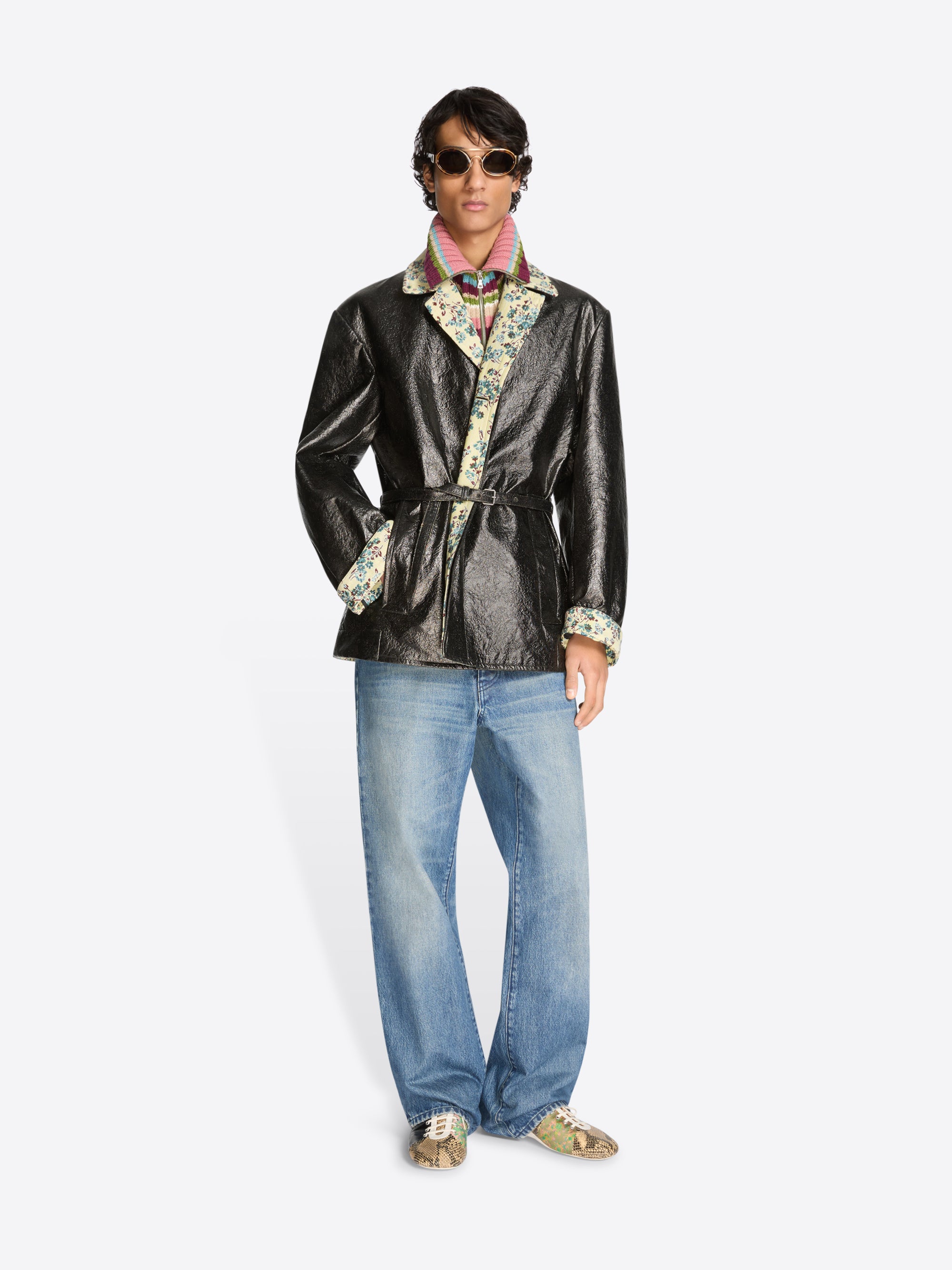 Person wearing a shiny black coat with floral patterns, blue jeans, and shoes on a white background
