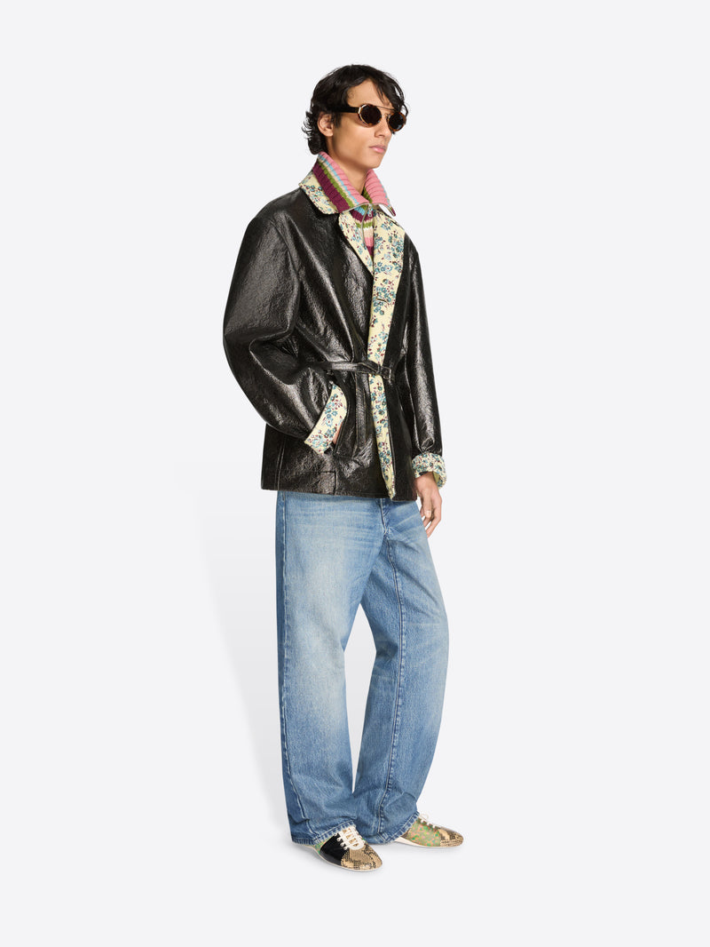 Person wearing a shiny black coat with floral patterns, blue jeans, and shoes on a white background