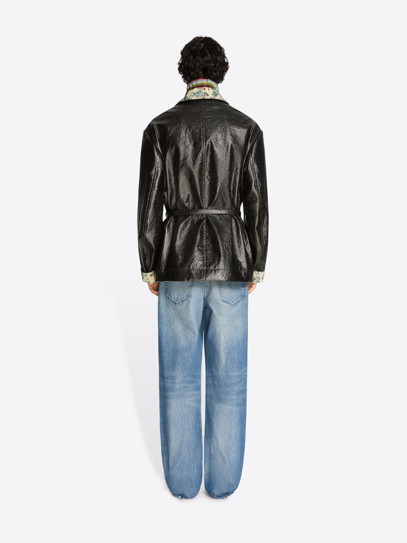 Person wearing a shiny black coat with floral patterns, blue jeans, and shoes on a white background