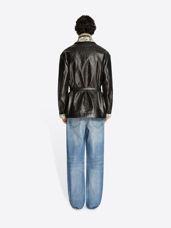 Person wearing a shiny black coat with floral patterns, blue jeans, and shoes on a white background