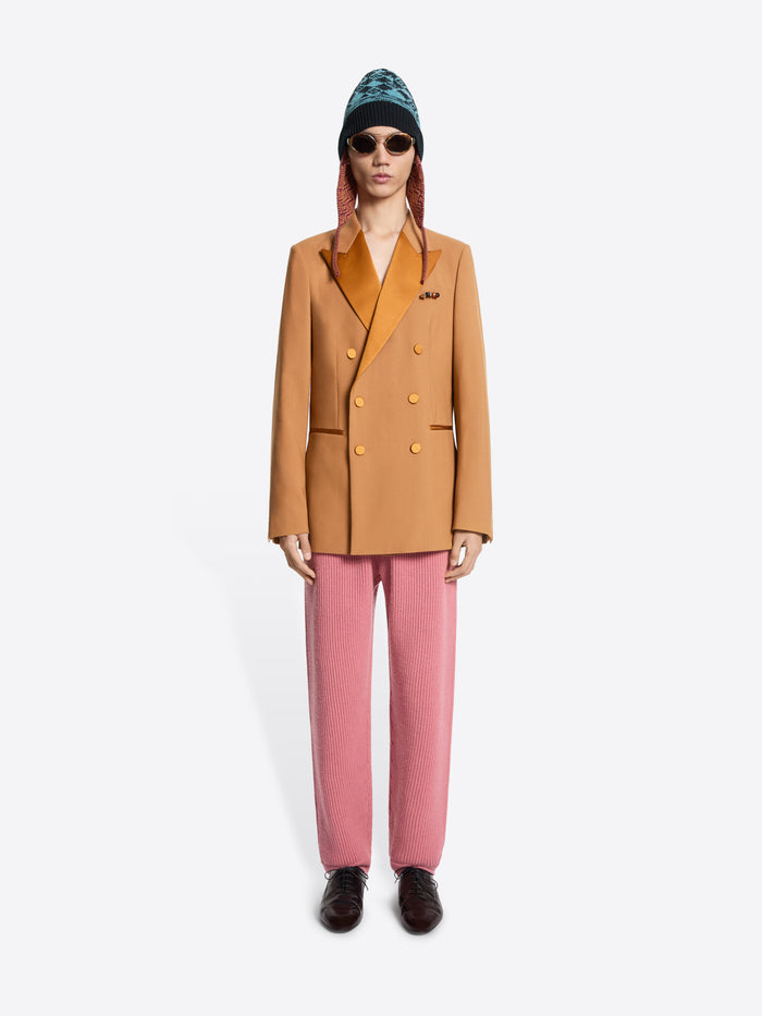 Person wearing an orange blazer, pink pants, and a patterned hat on a white background