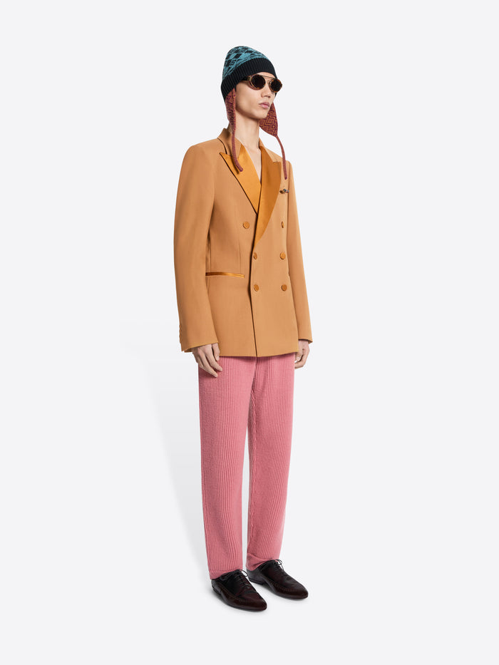 Person wearing an orange blazer, pink pants, and a patterned hat on a white background