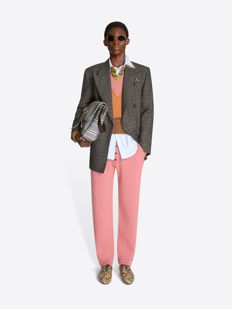 Person wearing a gray blazer, pink pants, and sunglasses holding a bag on a white background