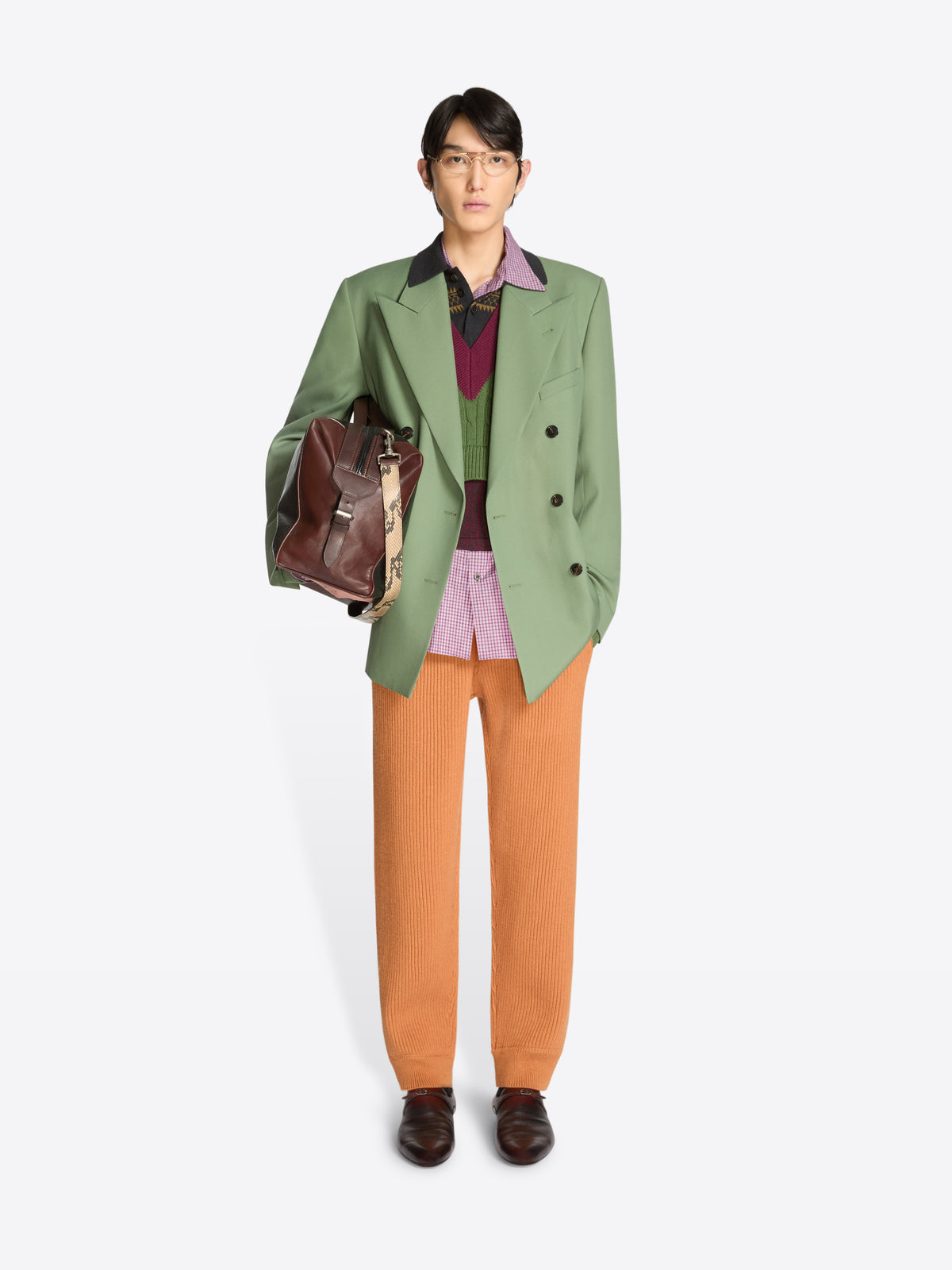 Person wearing a green blazer, orange pants, and holding a brown leather bag on a white background
