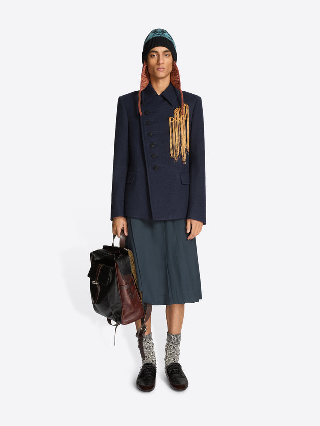Person wearing a dark coat, skirt, and black shoes holding a backpack on a white background