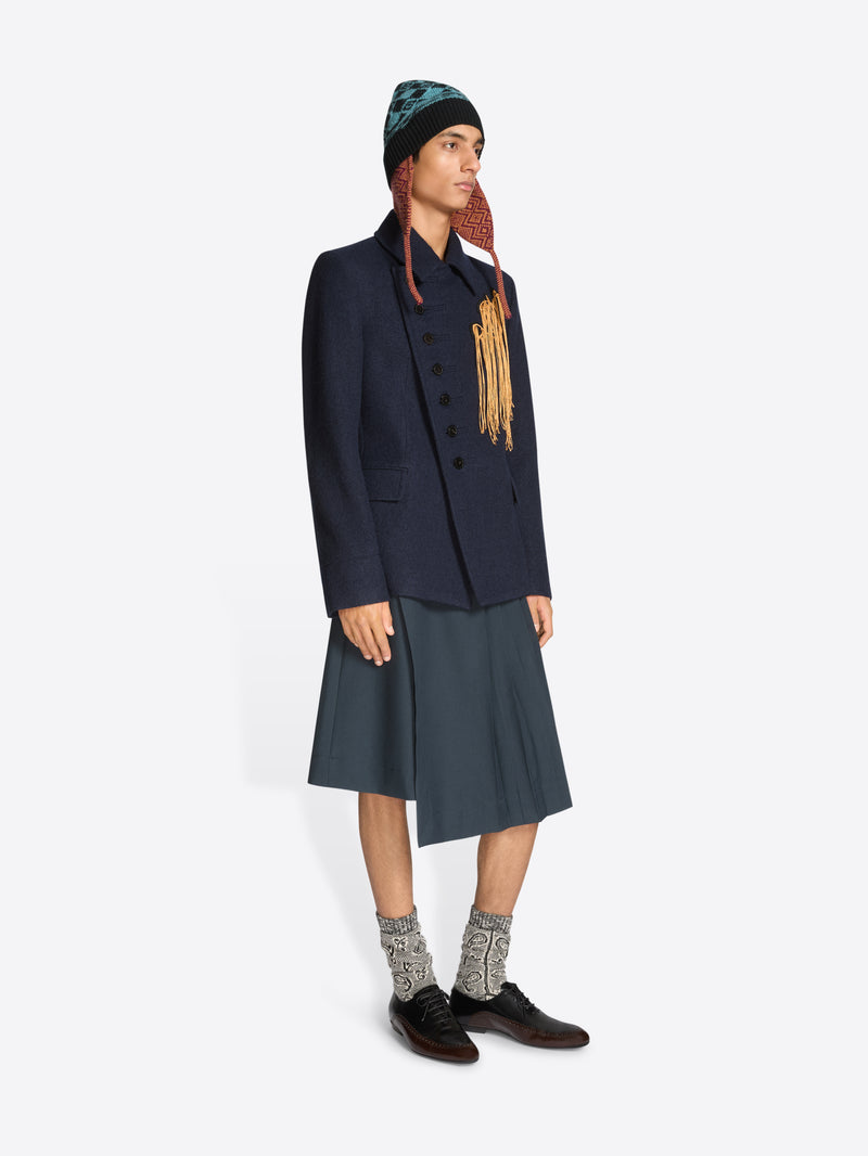 Person wearing a dark coat, skirt, patterned hat and black shoes on a white background
