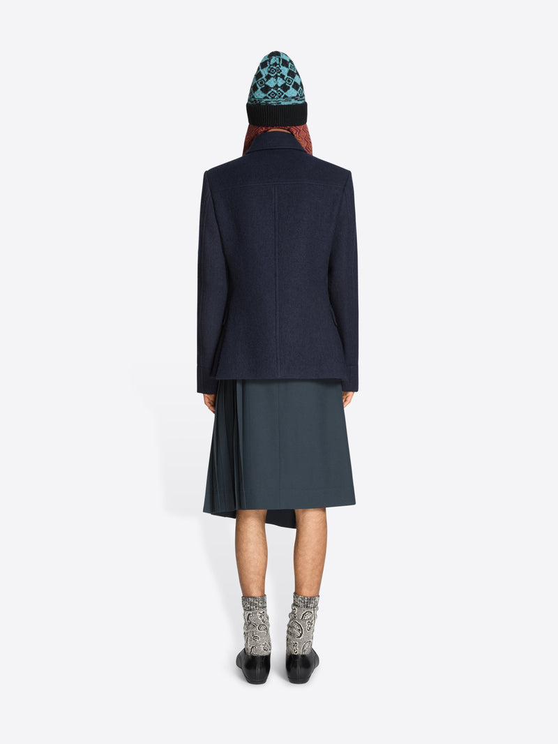 Back view of a person wearing a dark coat, skirt, patterned hat and black shoes on a white background