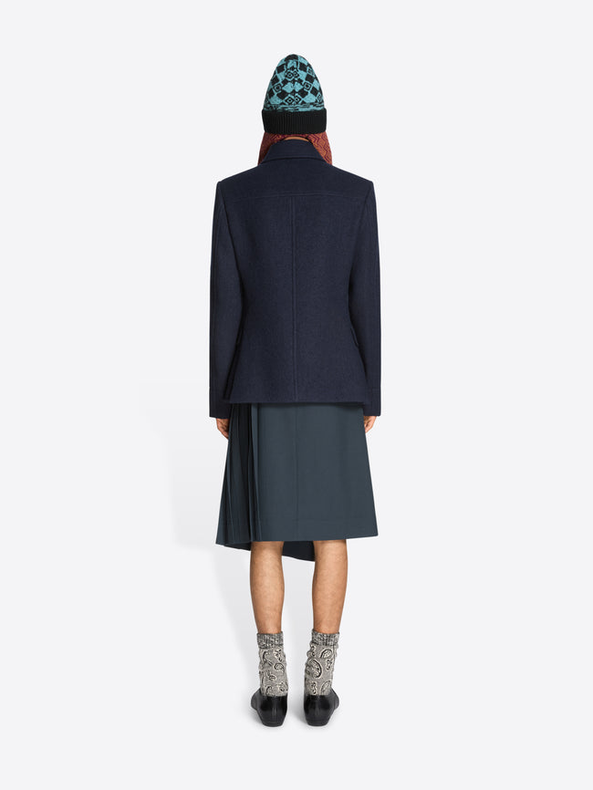 Back view of a person wearing a dark coat, skirt, patterned hat and black shoes on a white background