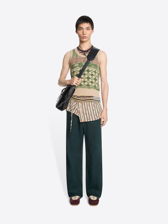 Model wearing a patterned sleeveless top,  dark pants and striped wrap belt with floral pattern with a black bag on a white background