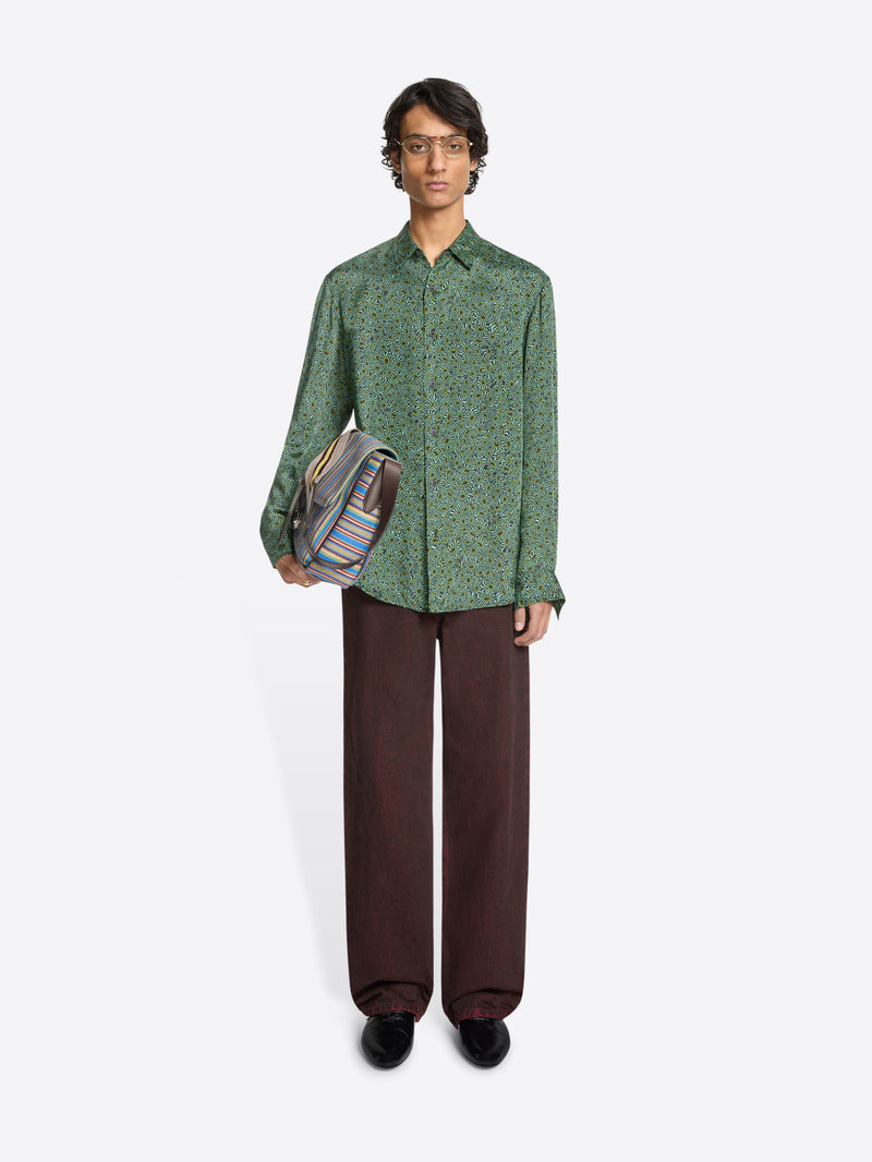 Person wearing a green long-sleeve button-up shirt, loose dark pants and a striped shoulder bag on a white background 