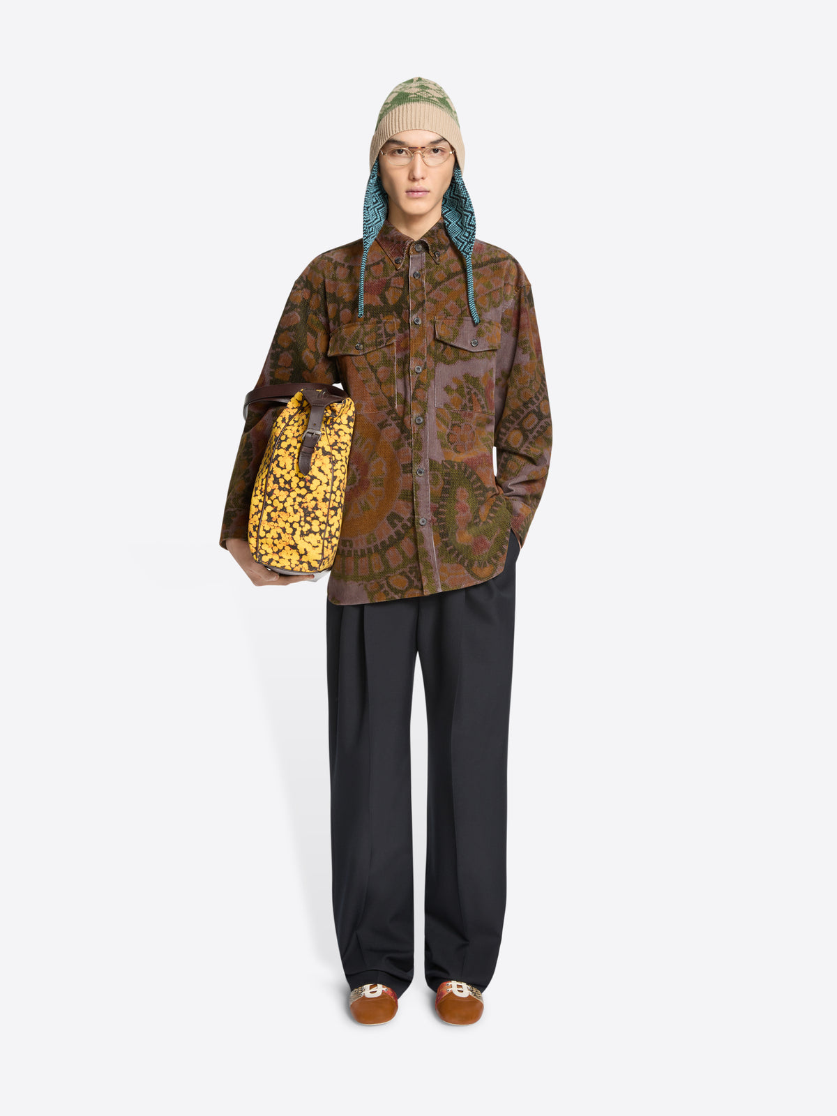 Person wearing a patterned shirt and black pants, holding a patterned bag on a white background