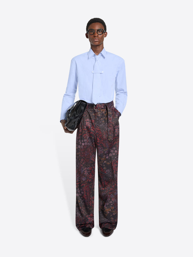 Person wearing a light blue shirt with a tie and patterned pants holding a black bag on a white background