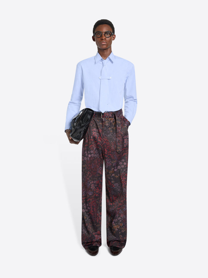 Person wearing a light blue shirt with a tie and patterned pants holding a black bag on a white background