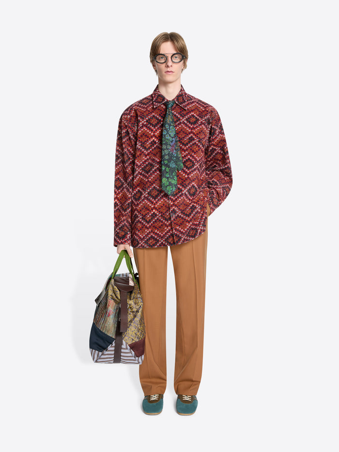 Person wearing a patterned shirt, orange pants and holding a colorful bag on a white background
