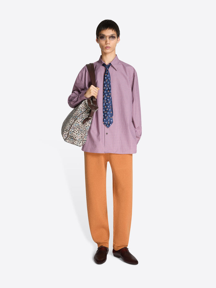 Person wearing a purple shirt, orange pants, and brown shoes holding a patterned bag on a white background