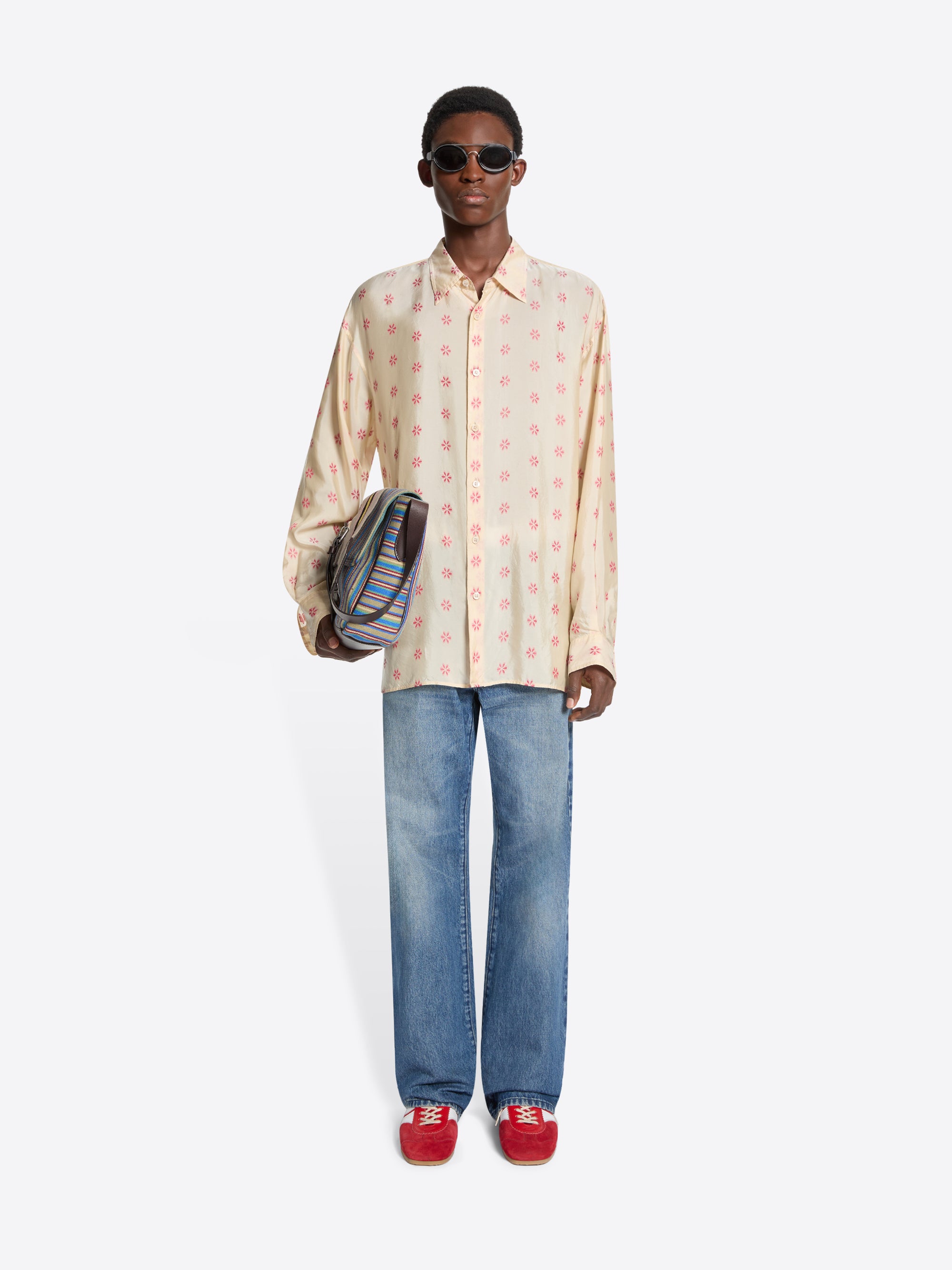 Person wearing a patterned shirt, jeans, and red shoes holding a striped bag on a white background