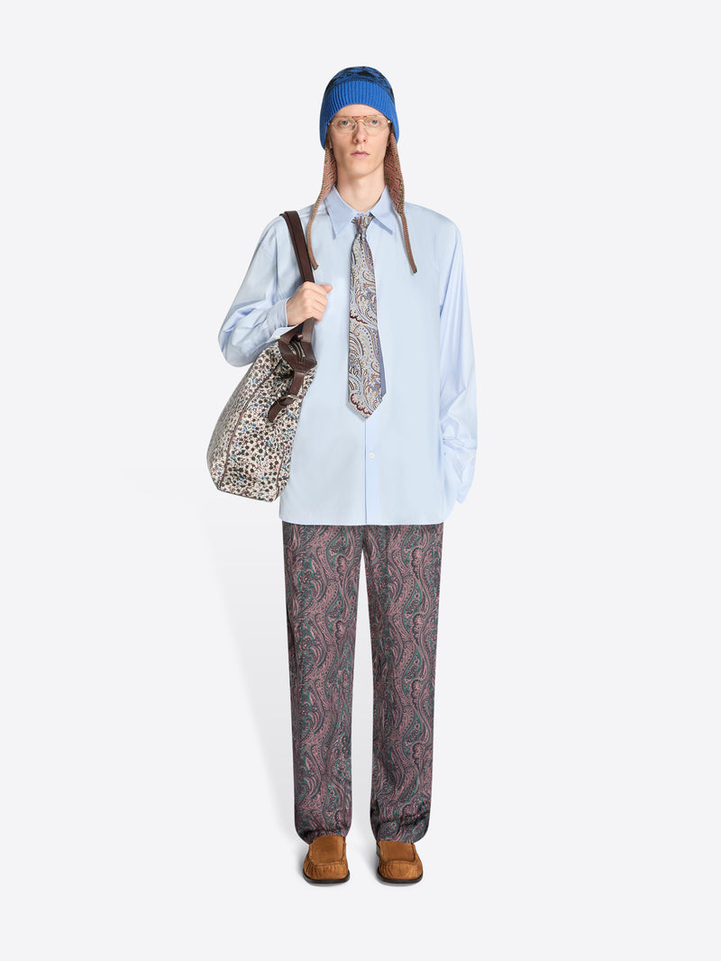 Person wearing a light blue shirt, patterned tie, patterned pants and a blue hat, holding a patterned bag on a white background.