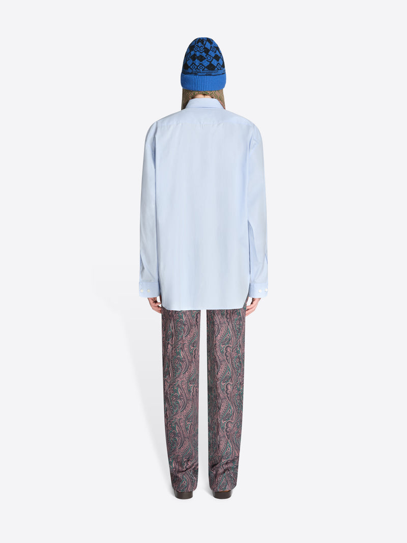 Back view of a person wearing a light blue shirt and patterned pants with a blue hat on a white background