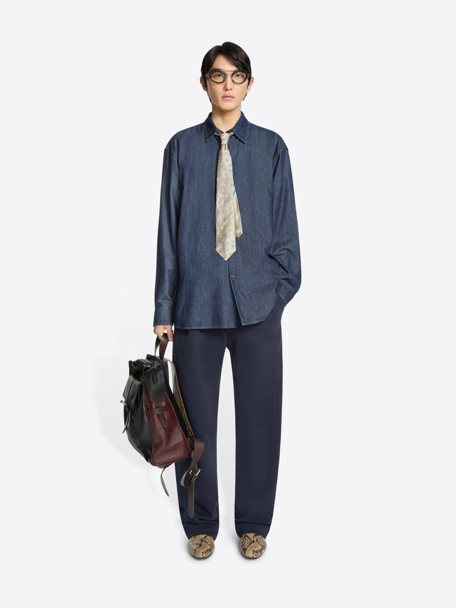 Person wearing a denim shirt and pants with a patterned tie, holding a leather backpack on a white background