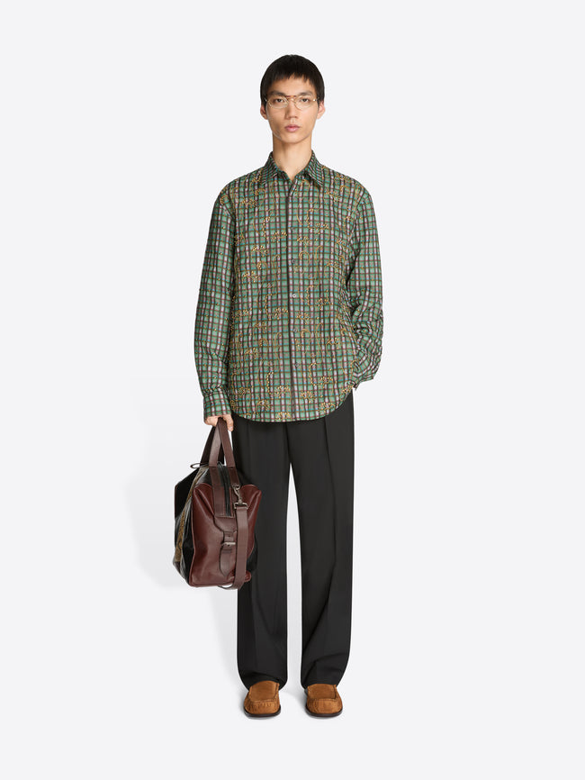 Man wearing a green plaid shirt and dark pants holding a brown leather bag on a white background