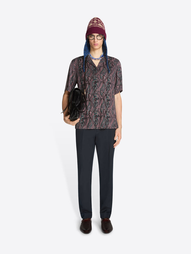 Person wearing a patterned shirt and dark pants holding a black bag on a white background