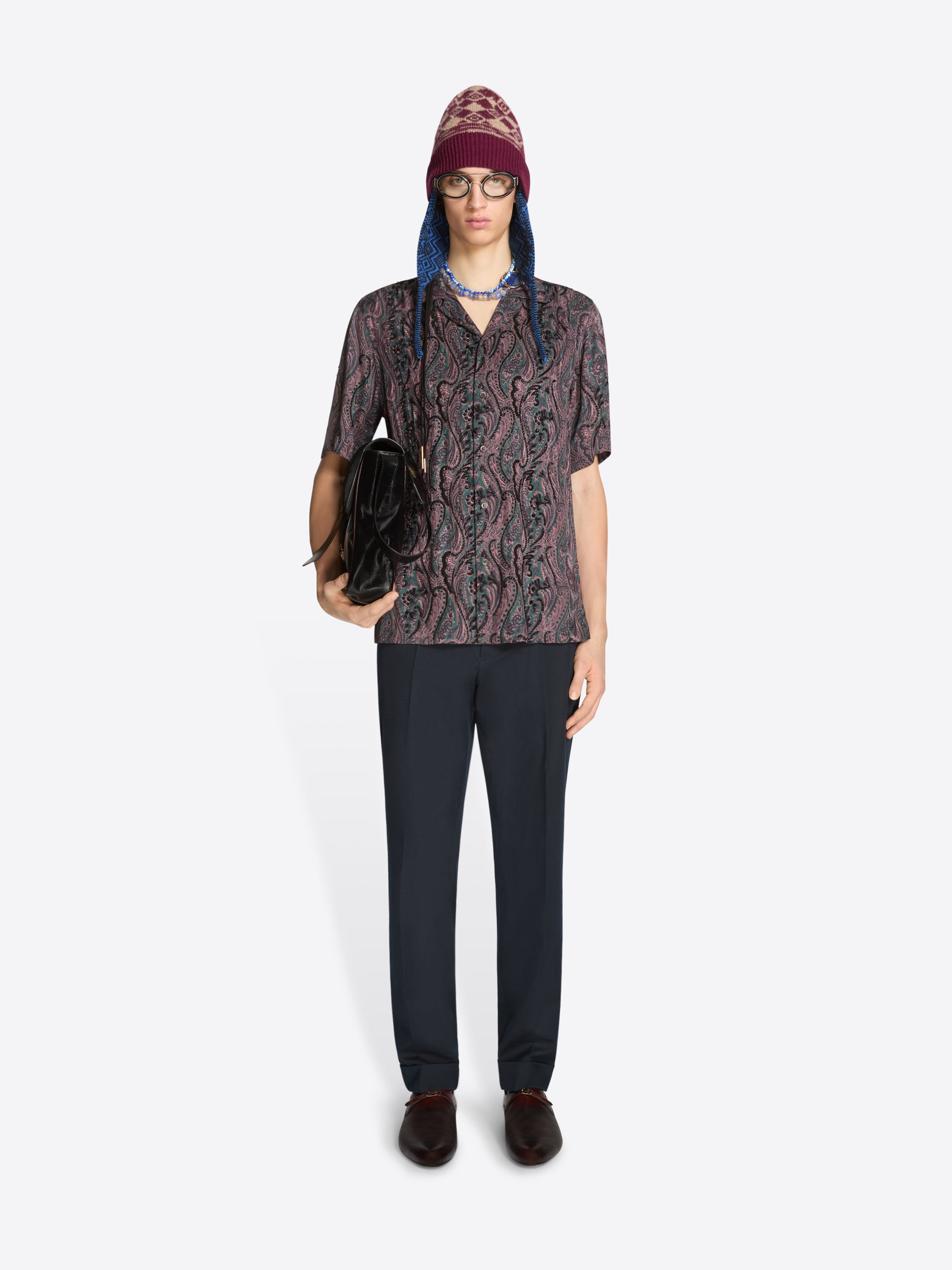 Person wearing a patterned shirt and dark pants holding a black bag on a white background