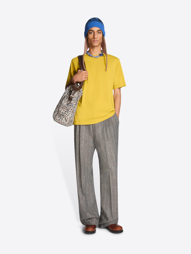 Person wearing a yellow t-shirt, gray pants, and a blue hat, holding a patterned bag on a white background