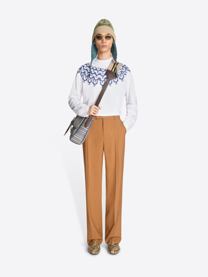 Person wearing a white long sleeve with blue pattern, camel-colored pants, and holding a bag on a white background