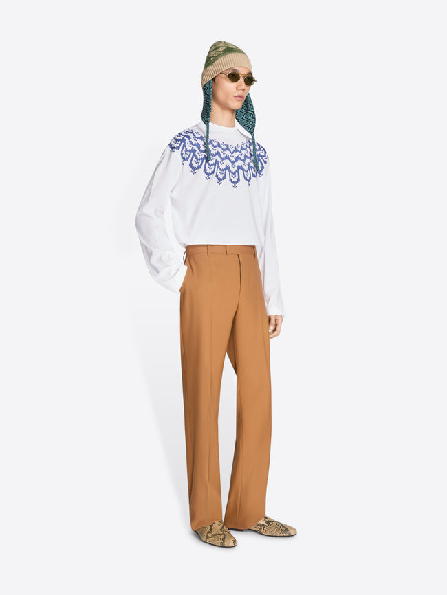 Person wearing a white long sleeve with blue pattern, camel-colored pants, hat and glasses 