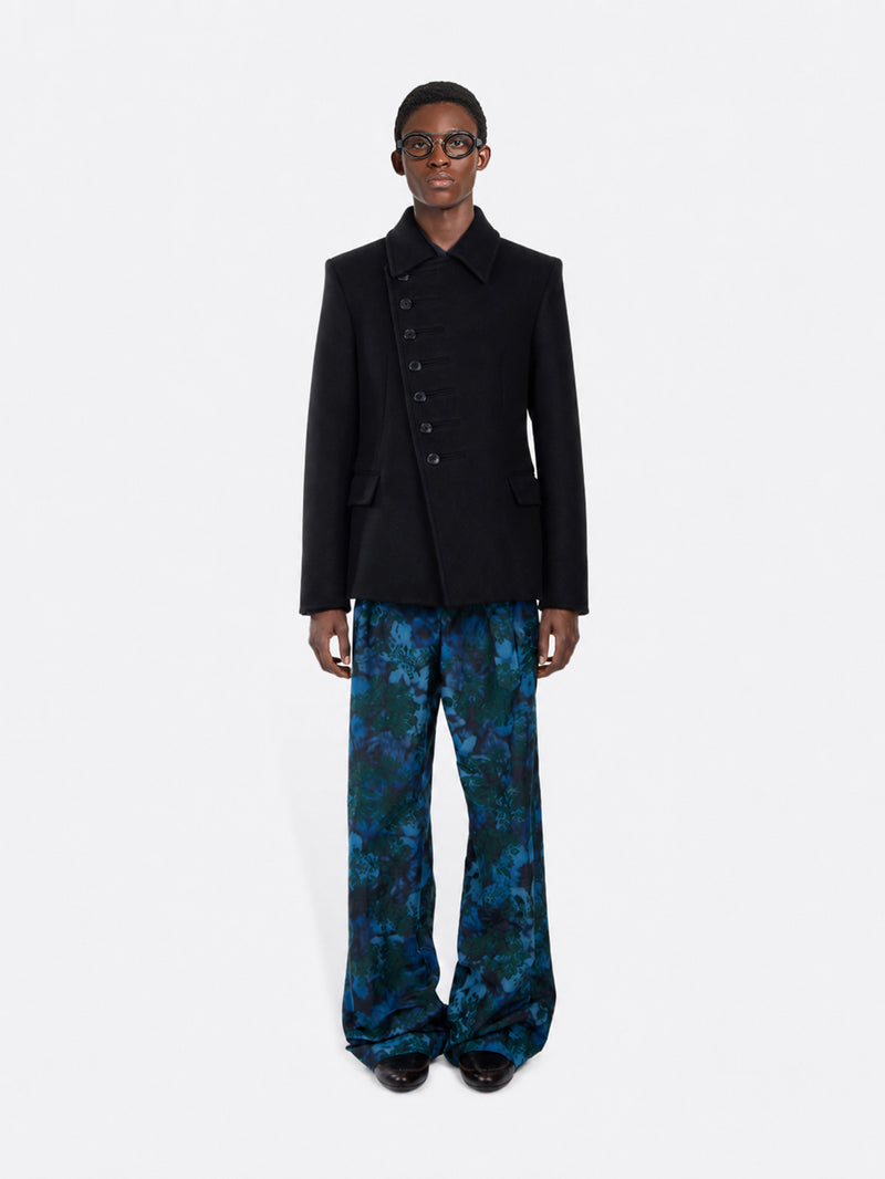 A model stands against a gray background wearing a black double-breasted blazer with decorative buttons down the front, paired with wide-leg pants featuring a blue and green abstract pattern, and black shoes.