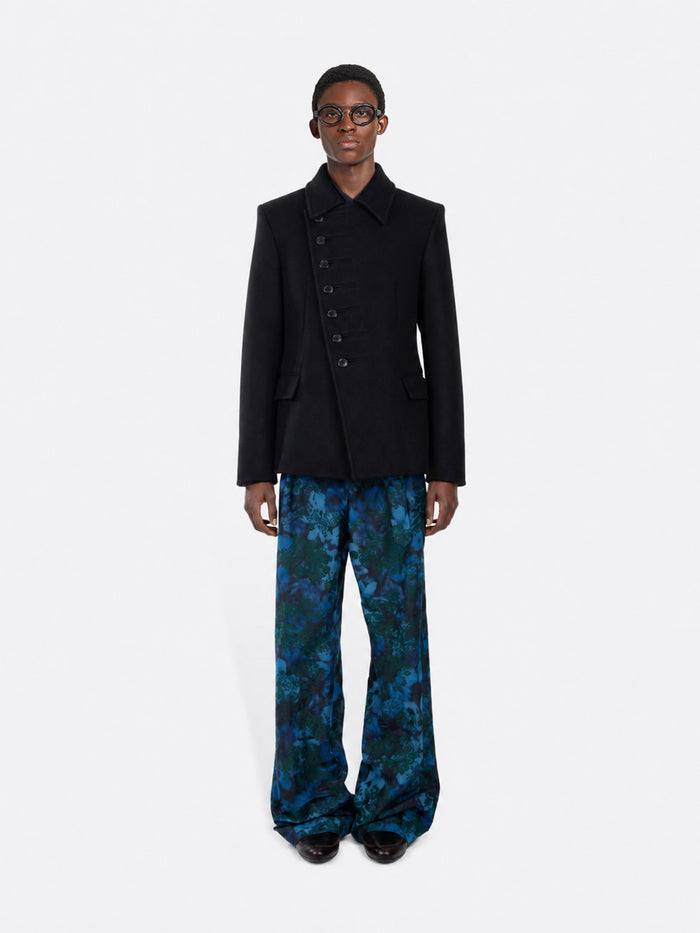 A model stands against a gray background wearing a black double-breasted blazer with decorative buttons down the front, paired with wide-leg pants featuring a blue and green abstract pattern, and black shoes.