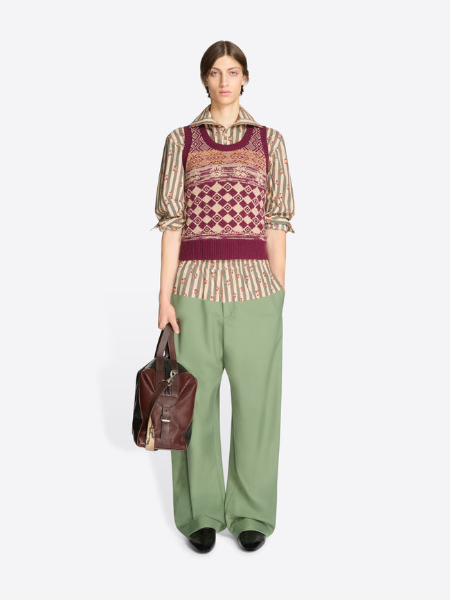 Person wearing a patterned top and green pants holding a brown bag on a white background