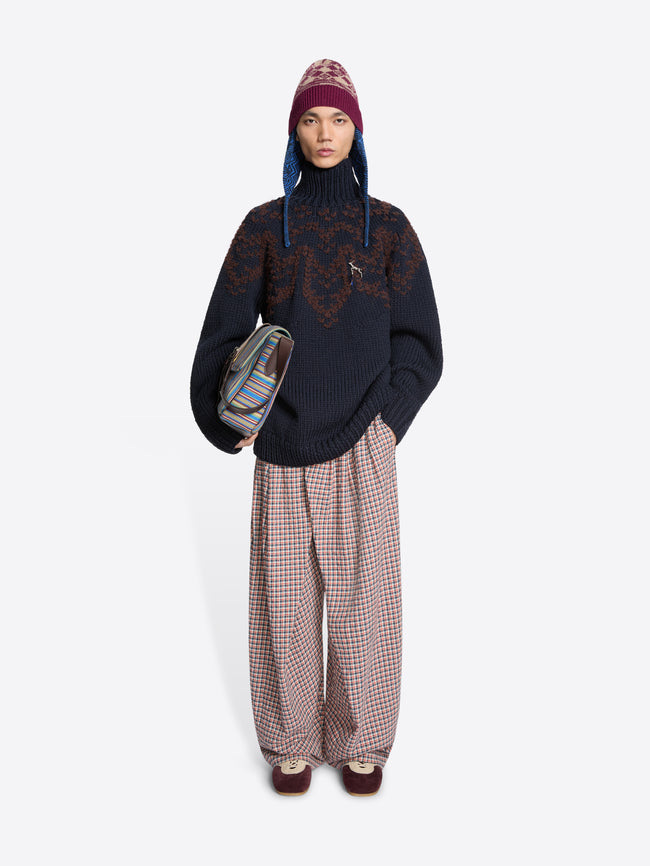 Person wearing a dark sweater with intricate patterns, plaid pants, and a patterned hat, holding a bag on a white background