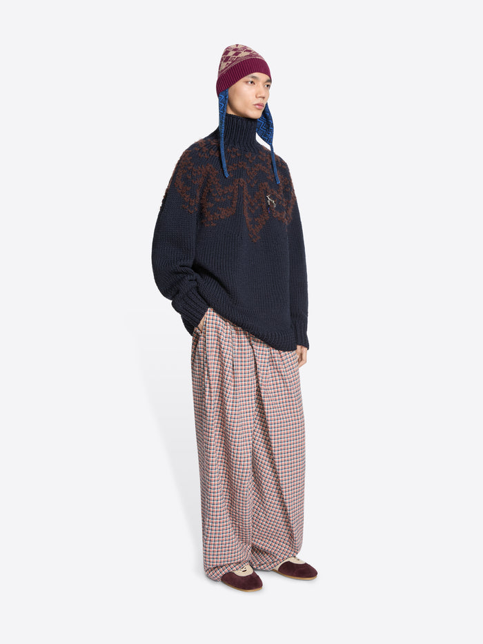 Person wearing a dark sweater with a pattern, plaid pants, and a patterned hat on a white background