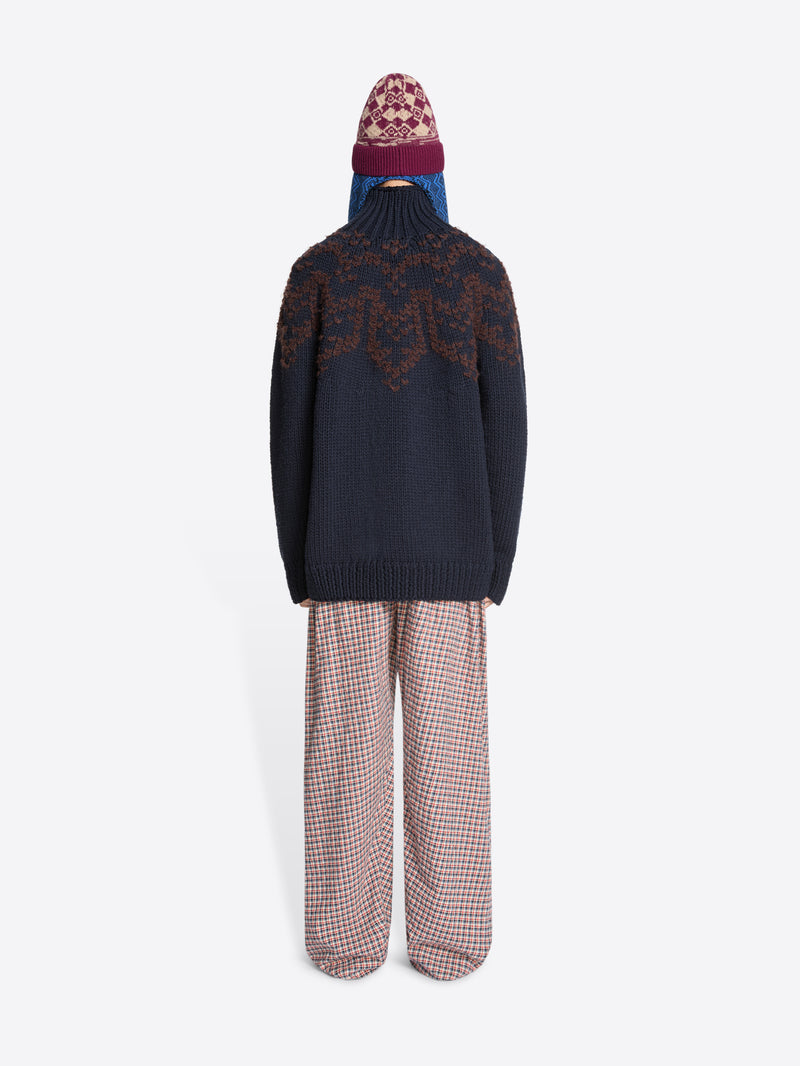 Back view of a person wearing a dark blue sweater with a patterned design, plaid pants, and a patterned hat on a white background