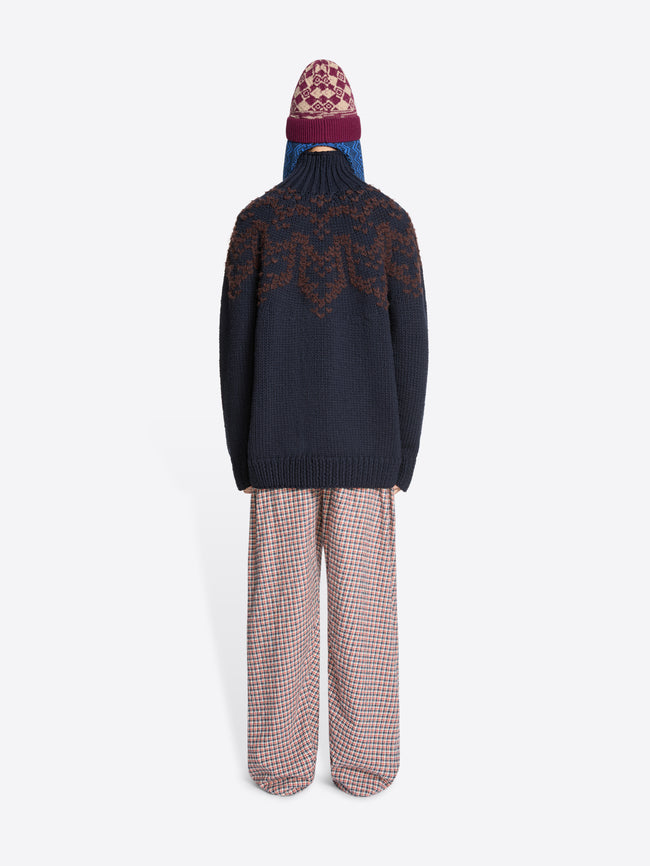 Back view of a person wearing a dark blue sweater with a patterned design, plaid pants, and a patterned hat on a white background