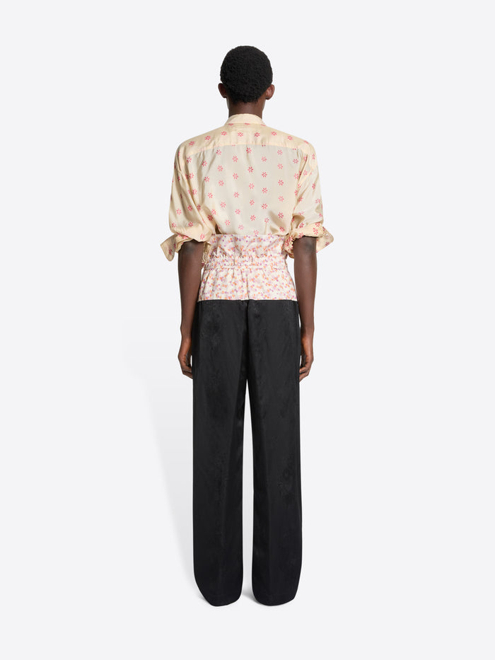 Back view of a person wearing a patterned shirt and black pants with a floral waistband on a white background