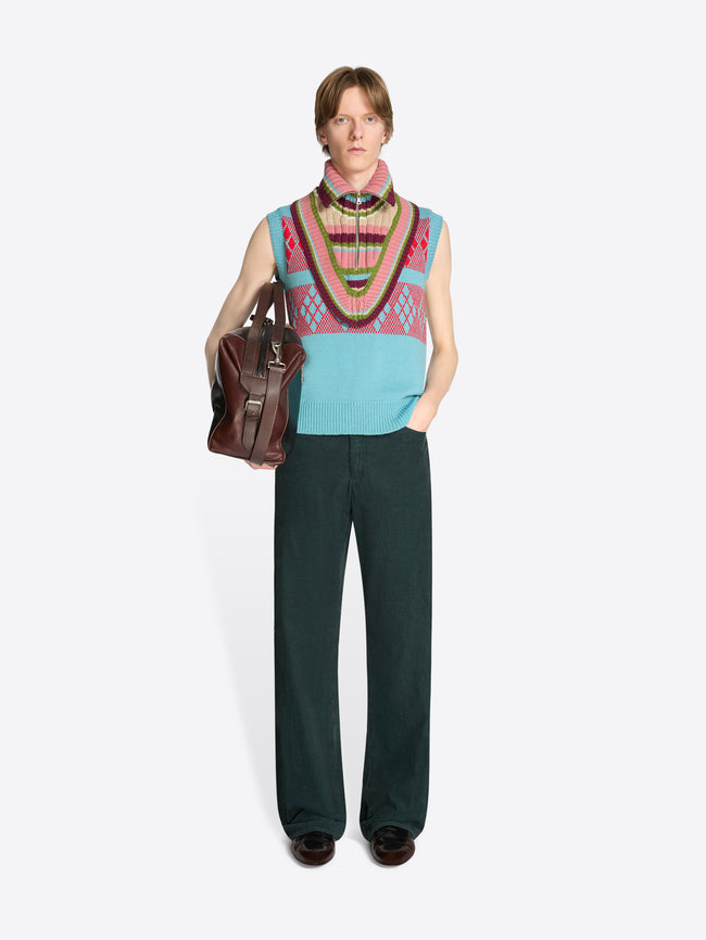 Person wearing a colorful sleeveless vest with a pattern, dark pants, and holding a brown bag on a white background