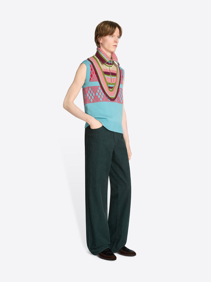 Person wearing a colorful sleeveless vest and dark green pants on a white background
