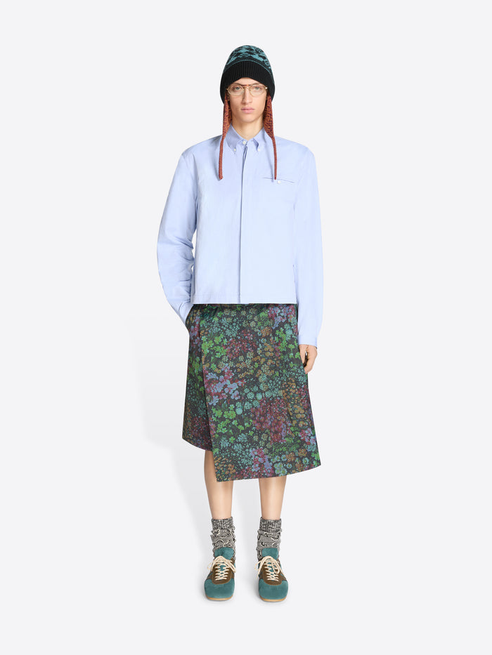 Person wearing a light blue shirt, patterned floral skirt, and green hat on a white background