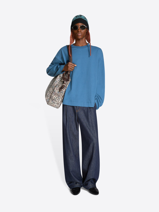 Person wearing a blue sweatshirt and dark jeans, holding a patterned bag on a white background