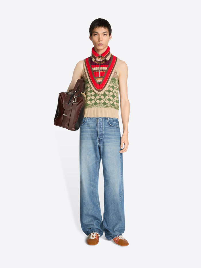Person wearing a patterned vest and blue jeans with a brown bag on a white background