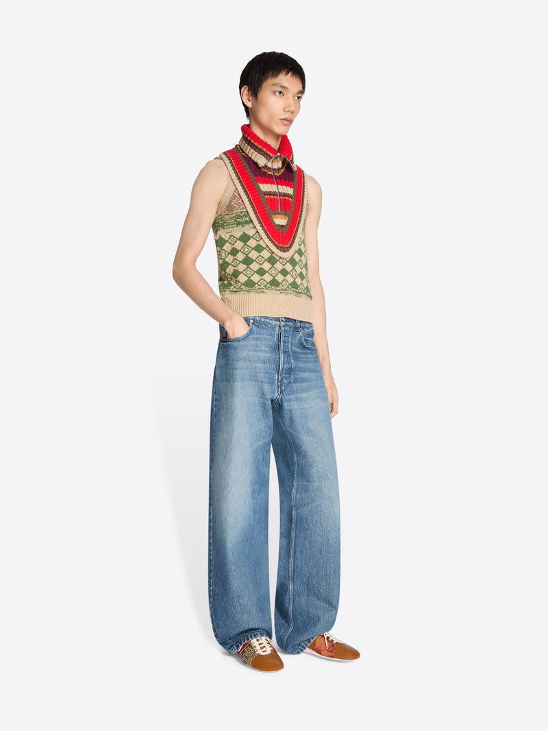 Person wearing a patterned sleeveless vest and blue jeans on a white background