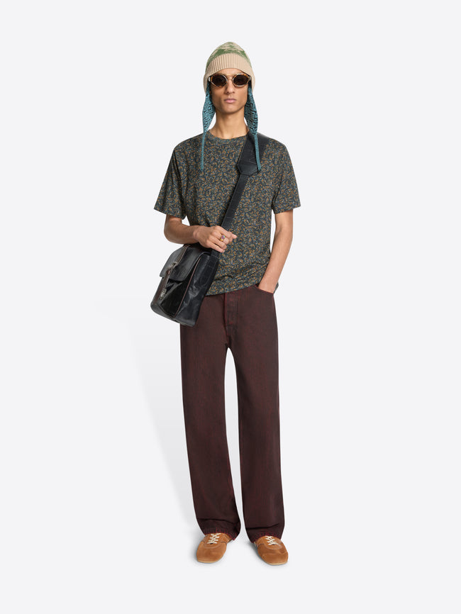 Person wearing a patterned t-shirt, red jeans, and a green hat with sunglasses, holding a black bag on a white background