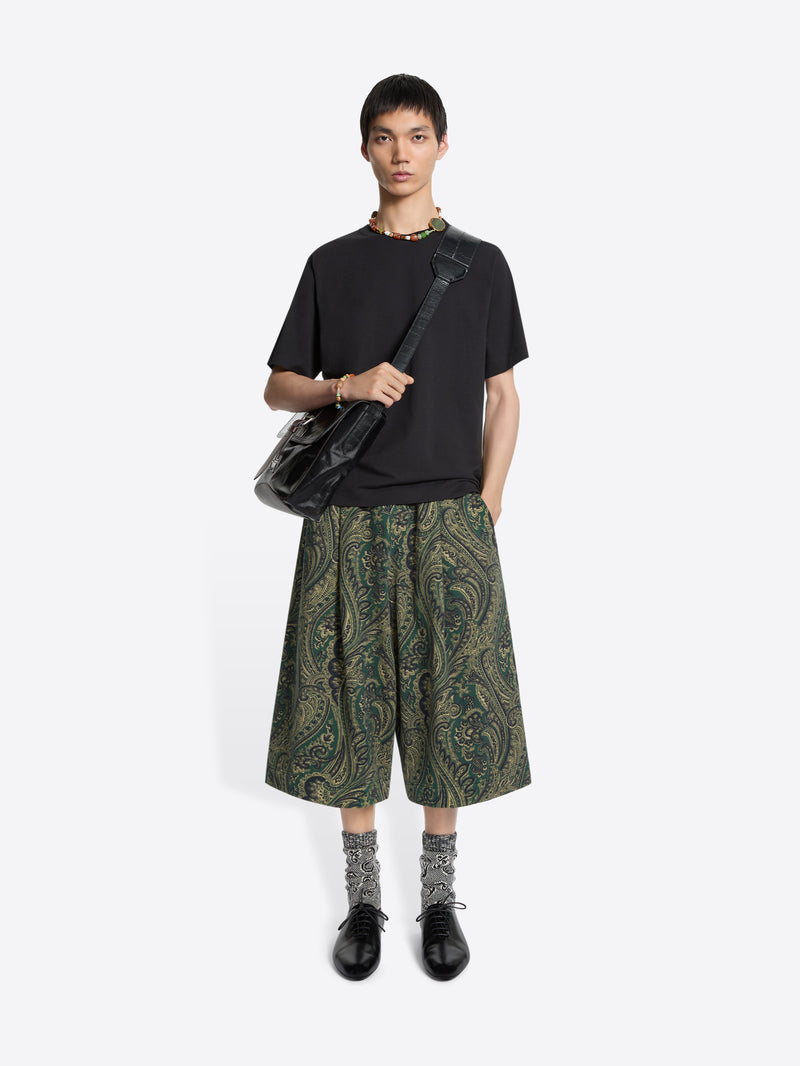 Person wearing a black t-shirt, green patterned shorts, and carrying a black bag on a white background